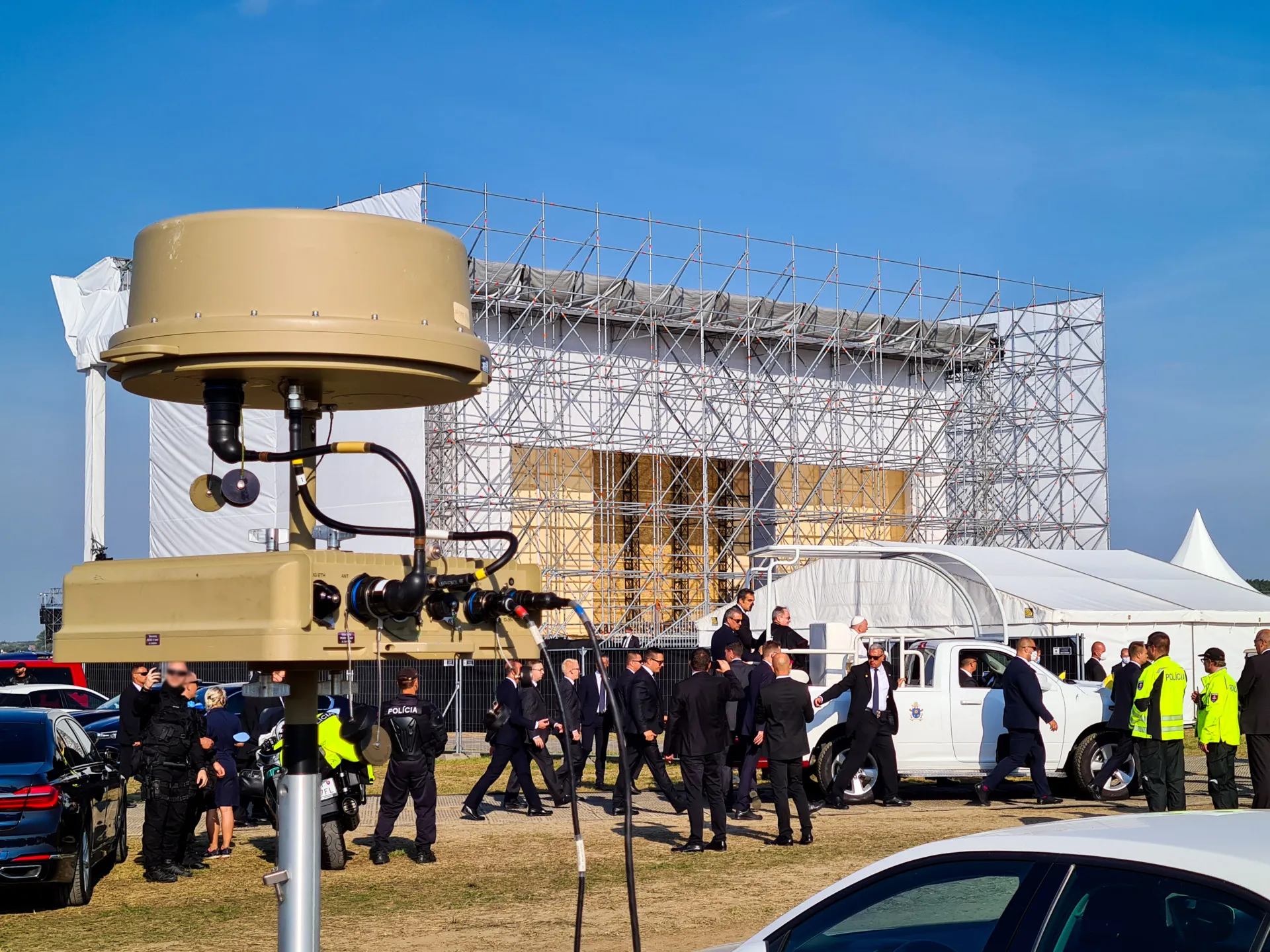 A security-focused event setup featuring a surveillance or communication device in the foreground, a crowd of suited officials and security personnel near vehicles, and a large structure under construction in the background.